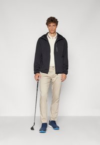 Svart lett jakke med hette, kremfarget polo, beige bukser og blå treningssko. Modellen holder en golf kølle og viser en avslappet utendørs stil.