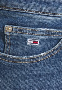 Blå denimtyg med en framficka med en synlig silverknapp och en logotyp patch med röda, vita och blå horisontella ränder.