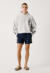 Sudadera con capucha gris con logotipos impresos, combinada con bañador azul marino con acentos amarillos. Sandalias deslizantes beige completan el look.