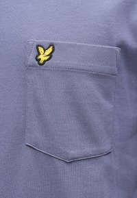 Camicia a maniche corte color viola chiaro con taschino sul petto, caratterizzata da un logo ricamato di un uccello nero e giallo. Tessuto in cotone liscio.
