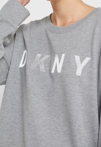 Osoba oblečená v šedé mikině se bílým vyšitým nápisem "DKNY" přes prsa, vystřižená v oblasti krku a se vztyčenou rukou.