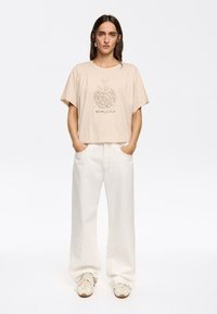 T-shirt cropped beige con una texture morbida, caratterizzato da una grafica floreale e testo del marchio centrato sul davanti. Abbinato a pantaloni bianchi larghi.