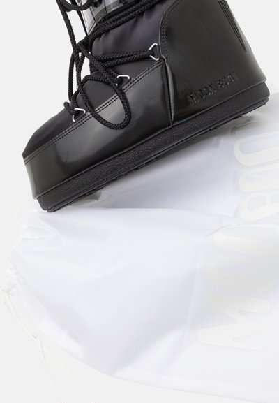 Black snow boot with a glossy finish, textured rubber sole, and black laces, resting on a white drawstring bag with a subtle logo.
