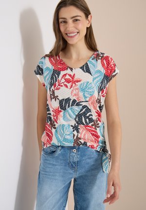 Kvinde smilende iført farverig blomstermønstret kortærmet top og blå jeans stående mod en beige baggrund