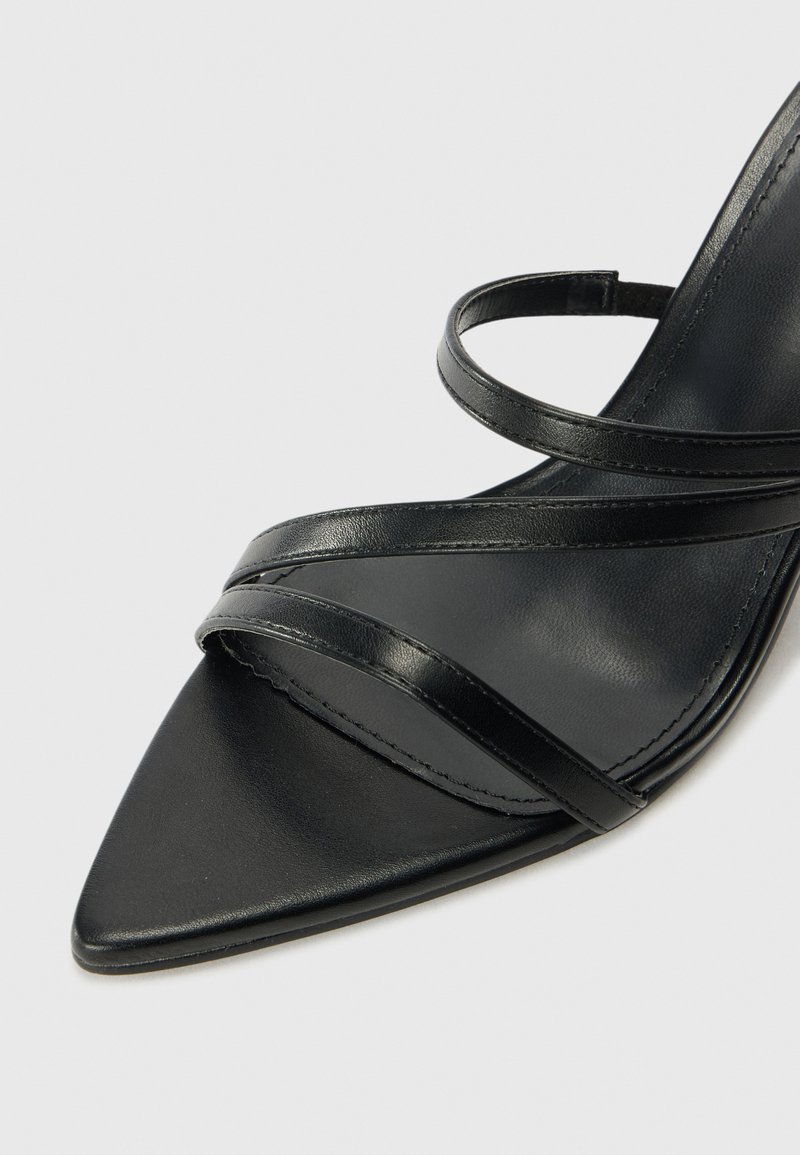 Sandale à talon en cuir noir avec bout pointu. Présente des fines sangles croisées sur le cou-de-pied et une semelle lisse au toucher.