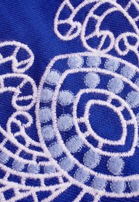 Gros plan sur un tissu bleu avec des motifs circulaires et en spirale blancs brodés de manière complexe, comprenant de petites formes pleines et en contour.