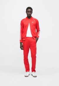 Fato de treino vermelho feito de tecido macio com riscas brancas nas laterais, composto por um casaco com fecho de correr e calças a condizer. Tênis brancos simples.