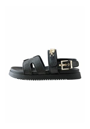 Black flat sandal with textured straps, gold buckle, and small gold charm on ankle strap, featuring a ridged sole.