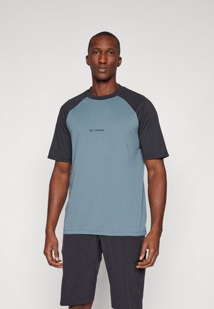 Man draagt een blauw-zwart shirt met korte mouwen en een zwarte korte broek, staand tegen een effen lichte achtergrond.