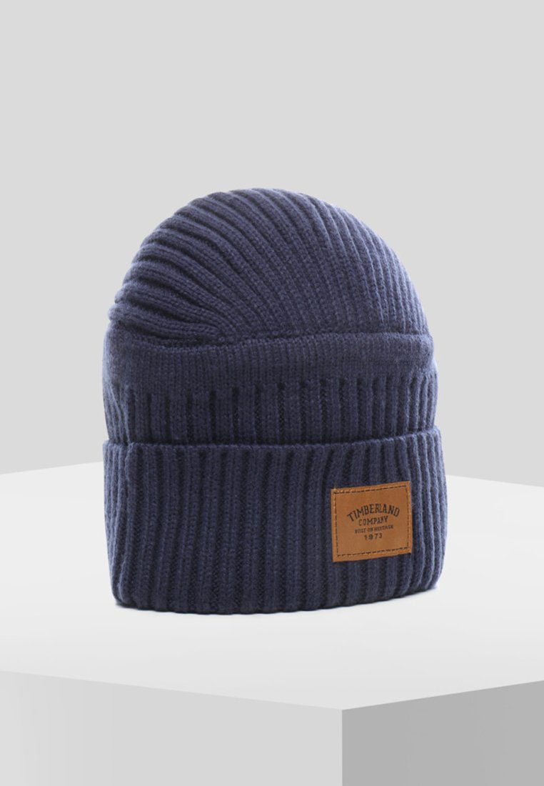 Berretto in maglia blu navy con texture a coste verticali e risvolto. Presenta una patch in pelle marrone con il logo Timberland.