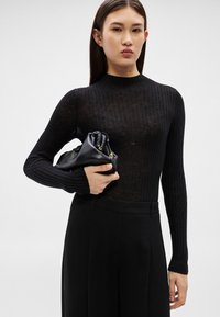 Sieviete, kas tērpusies melnā, ribotā, ar garām piedurknēm topā un melnās biksēs, turot salocītu melnu ādas rokassomiņu zem elkoņa.