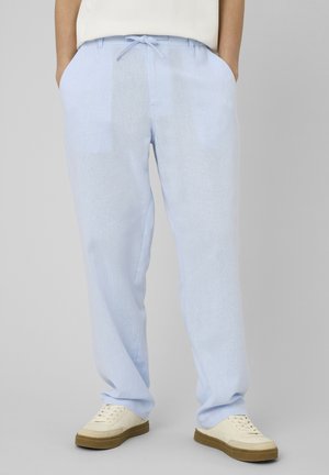 Personne portant un pantalon bleu clair à cordon et des baskets blanches avec semelles marron, mains dans les poches, debout devant un fond uni.