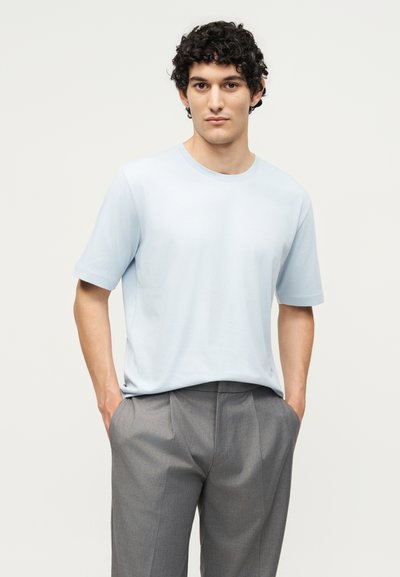 Άνδρας με κατσαρά μαλλιά στέκεται με τα χέρια στις τσέπες, φορώντας ανοιχτό μπλε κοντομάνικο πουκάμισο και γκρι πλισέ παντελόνι σε μονόχρωμο φόντο.