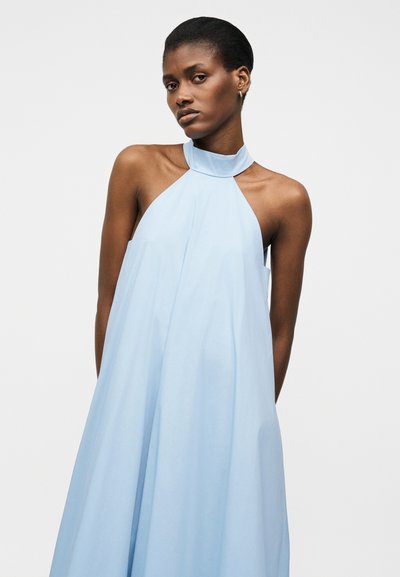 Femme aux cheveux courts portant une robe fluide bleu clair sans manches à col montant sur un fond uni.