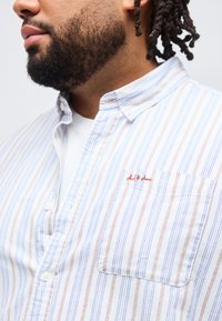 Άνδρας με γενειάδα και dreadlocks που φοράει λευκό πουκάμισο με γιακά, με κατακόρυφες ρίγες σε μπλε και πορτοκαλί χρώμα και τσέπη στο στήθος.