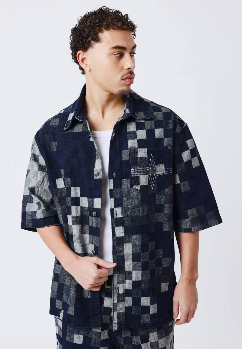 Homme aux cheveux bouclés portant une chemise à manches courtes à carreaux bleu marine et gris sur un t-shirt blanc, regardant vers sa gauche.