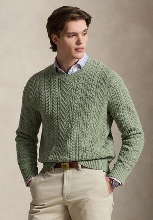 Joven con un jersey verde de punto trenzado sobre una camisa con cuello, pantalones beige, cinturón marrón y reloj de pulsera, mirando de lado con las manos en los bolsillos.