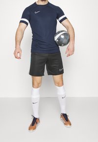 Laineen urheilupaita, jossa on valkoiset raidat hihoissa, mustat shortsit, valkoiset sukat ja ruskeat futikset. Pelaaja pitää harmaata jalkapalloa.