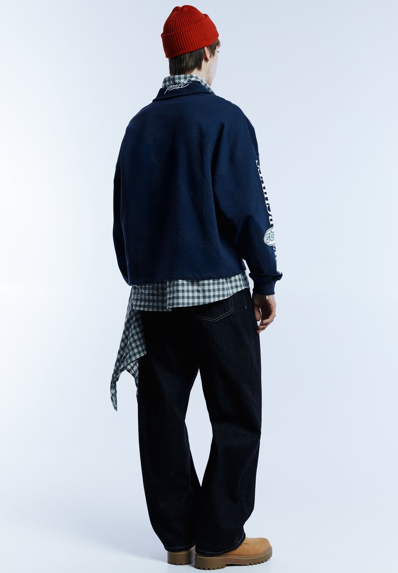 Felpa oversize blu navy con dettagli ricamati, indossata sopra una camicia a quadri verde; abbinata a jeans neri a gamba larga e stivali color cuoio.