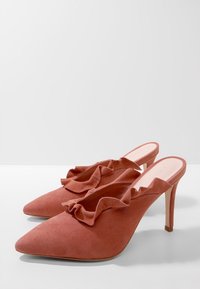 Ein Paar hochhackige Mules aus staubrosafarbenem Wildleder mit spitzen Zehen und gerafftem Rand entlang der Öffnung, auf einer weißen Oberfläche präsentiert.