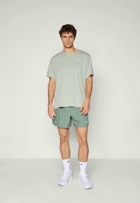 Un jeune homme debout portant un t-shirt Nike gris clair, un short vert assorti, des chaussettes Nike blanches et des chaussures de sport blanches sur un fond uni.