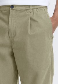 Pantalon vert olive clair en tissu lisse. Il présente une fermeture à boutons, des poches latérales et une taille élastique pour plus de confort.