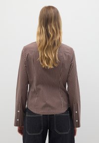 Braun gestreiftes Hemd mit langen Ärmeln, das weiße Knopfakzente und einen abgerundeten Saum aufweist, kombiniert mit dunklen Jeans.