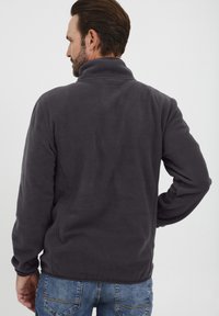 Pelēka vilnas jaka ar augstu apkakli, kurai ir gluda virsma un brīvs piegriezums. Aizmugurē nav redzamu logo vai rakstu.