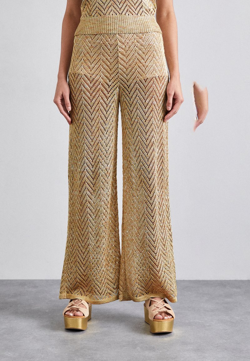 Pantalons larges dorés avec un motif en chevrons, fabriqués en tricot métallisé texturé. Associés à des chaussures à plateforme de couleur claire.
