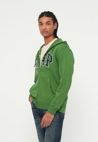 Sweatshirt verde com fecho à frente, bolso frontal, letras em azul-marinho e capuz com cordão, usado sobre uma t-shirt de cor clara e combinado com jeans escuros.