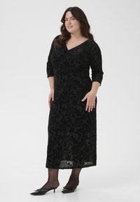 Fekete ruha virágos textúrájú mintázattal, háromnegyedes ujjakkal és V-kivágással, áttetsző fekete harisnyával és magassarkú cipővel viselve.