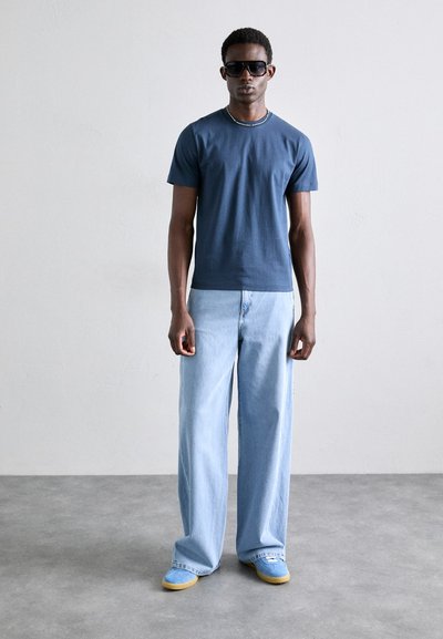T-shirt bleu marine à manches courtes associé à un jean large bleu clair et des baskets bleues avec des semelles jaunes. Le modèle porte des lunettes de soleil noires.