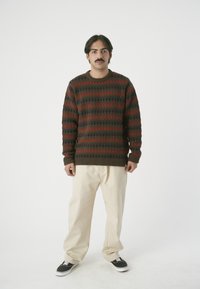 Dunkelgrüner Strickpullover mit roten und schwarzen geometrischen Mustern, Rundhalsausschnitt und gerippten Bündchen. Kombiniert mit hellbeigen, locker sitzenden Hosen und schwarzen Sneakers.