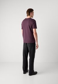 Tricou purpuriu cu mânecă scurtă, din bumbac, cu guler rotund, purtat cu blugi negri cu talie înaltă, stând în fața unui fundal gri deschis.