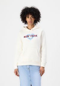 Kremgenser med frontlomme, med rød "NEW YORK" tekst og blå hjertegrafikk. Sammen med et lyst denimskjørt, har modellen krøllete hår.