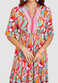 Un vestido colorido con un patrón floral en rosa, naranja, verde y azul. Mangas con volantes y una cintura fruncida con detalles de pompones y bordados.