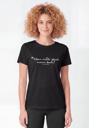 Junge Frau mit lockigem Haar, die ein schwarzes T-Shirt mit deutschem Text ""Wenn nicht jetzt, wann dann?"" in weißen Buchstaben trägt.