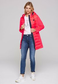 Bright pink puffer jacket with a hood, zip fastening, and contrasting navy lining, worn over a white graphic tee, paired with blue jeans and white sneakers.