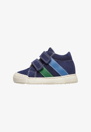 Baskets en daim marine avec deux bandes Velcro, rayures vertes et bleues, semelle en caoutchouc blanche et détails texturés le long du bord.