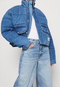 Krótka puchowa kurtka jeansowa w jasnoniebieskim kolorze z wysokim kołnierzem, zapinana na guziki, z dwoma przednimi kieszeniami; noszona z dopasowanymi jeansami z wysokim stanem.