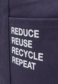 Granatowa tkanina o teksturowej powierzchni z dużymi białymi literami: "REDUCE REUSE RECYCLE REPEAT" na kieszeni.