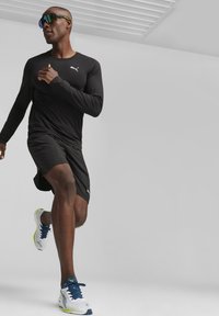 Schwarzes langärmliges Sportshirt mit weißem Logo, kombiniert mit schwarzen Shorts. Weiße und blaue Laufschuhe mit grünem Akzent.
