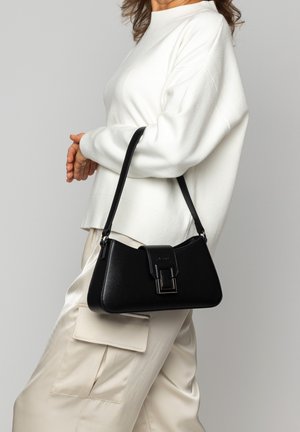 Schwarze Lederhandtasche mit strukturierter Form, kurzen Trägern und silbernem Verschlussdetail, begleitet von einer Person in einem weißen Pullover und beigen Hosen.