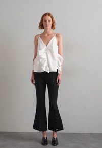 Valkoinen hihaton toppi, jossa on röyhelöyksitys yhdellä puolella, yhdistettynä mustiin leveälahkeisiin pantalooniin, joissa on leikkaukset helmassa, ja teräväkärkisiin mustiin kenkiin.