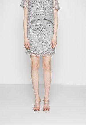 Persona che indossa un top a maniche corte in paillettes argento e una gonna abbinata con tacchi a mulo trasparenti e open-toe, in piedi davanti a uno sfondo bianco uniforme.