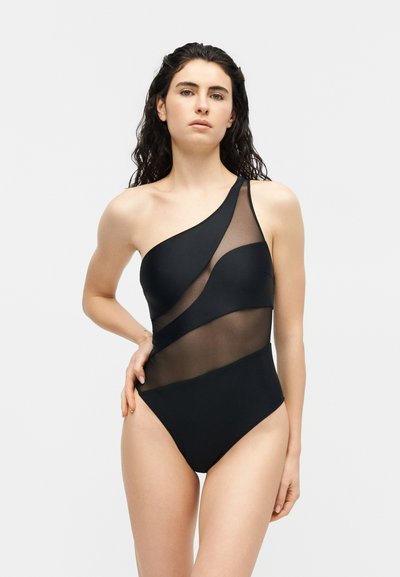 Femme aux cheveux bouclés et foncés portant un maillot de bain noir une épaule avec des panneaux diagonaux en mesh transparent, posant avec une main sur la hanche.