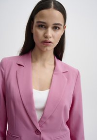 Růžový blazer s klopami, zapínání na jeden knoflík a bočními kapsami, nošený přes bílý ribbovaný top. Hladká textura, přiléhavý střih.