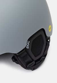 Pelēks slēpošanas ķivere ar MIPS tehnoloģiju. Tam ir gluda ārējā virsma, melni akcenti, regulējami siksnas un ventilācijas režģis.
