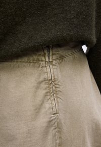 Κοντινή λήψη μπεζ παντελονιού με λεπτομέρεια πλαϊνού φερμουάρ, σε συνδυασμό με σκούρο πράσινο πλεκτό πουλόβερ στη μέση.