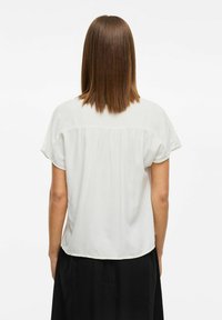 Blusa bianca a maniche corte con una vestibilità rilassata, caratterizzata da un design con schiena arricciata, texture liscia e orlo leggermente curvo.